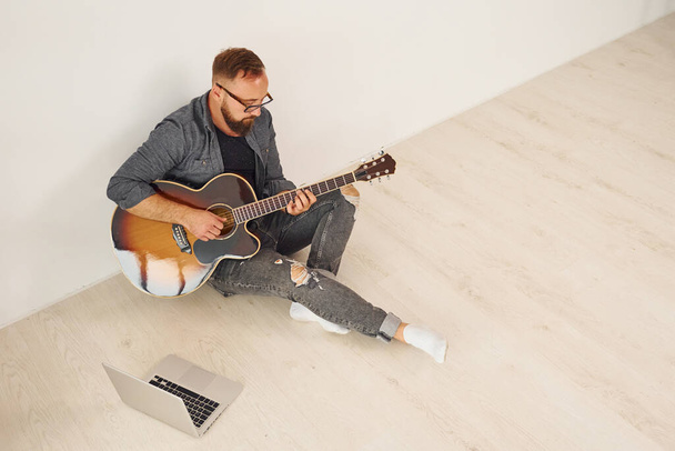 With laptop. Man in casual clothes and with acoustic guitar is indoors. - Photo, Image
