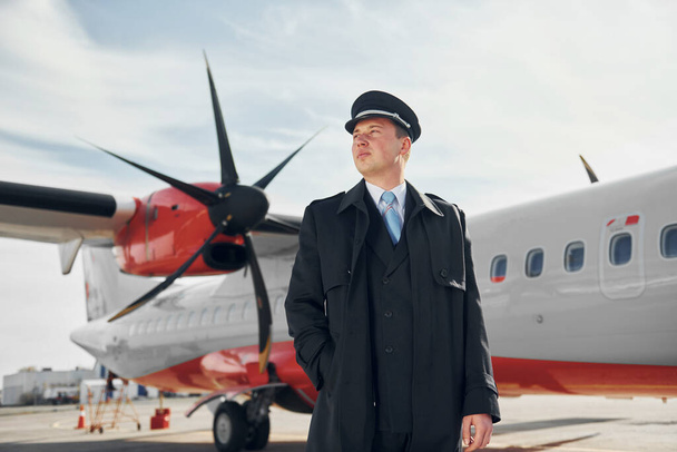Looking far away. Pilot in formal black uniform is standing outdoors near plane. - Фото, зображення