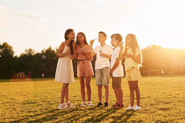 Iluminado por la luz del sol. De pie en el campo deportivo. Grupo de niños felices está al aire libre durante el día. - Foto, imagen