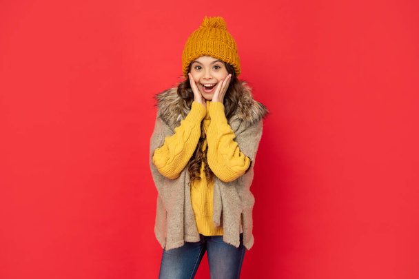 zaskoczona nastolatka dziewczyna w dzianinie czapka zimowa na czerwonym tle, dzianina. - Zdjęcie, obraz