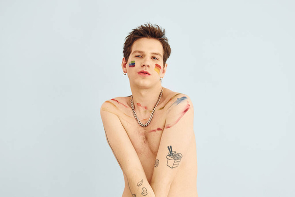 Young gay man is standing in the studio and posing for a camera. Shirtless guy with rainbow make up. - Φωτογραφία, εικόνα