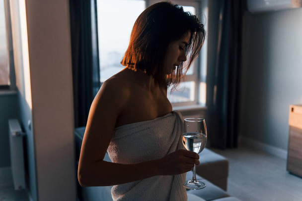 Standing at sunrise time in white towel. Young woman is indoors in room of modern house at daytime. - Foto, Bild