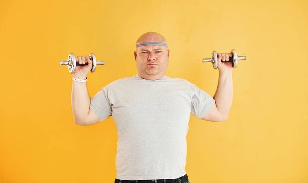 Exercises by using dumbbells. Funny overweight man in sportive head tie is against yellow background. - Fotografie, Obrázek