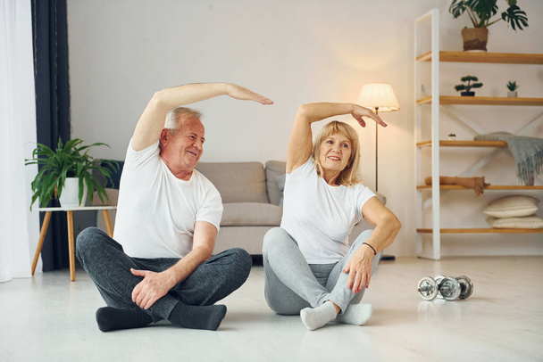 Sitting and doing exercises. Senior man and woman is together at home. - Fotoğraf, Görsel