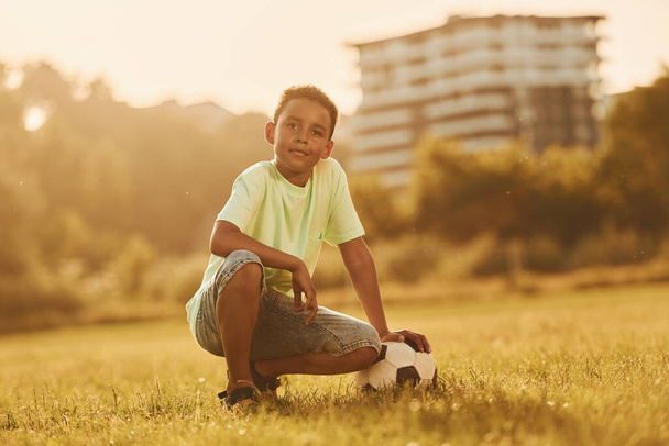 Taking a break. With soccer ball. African american kid have fun in the field at summer daytime. - Фото, изображение