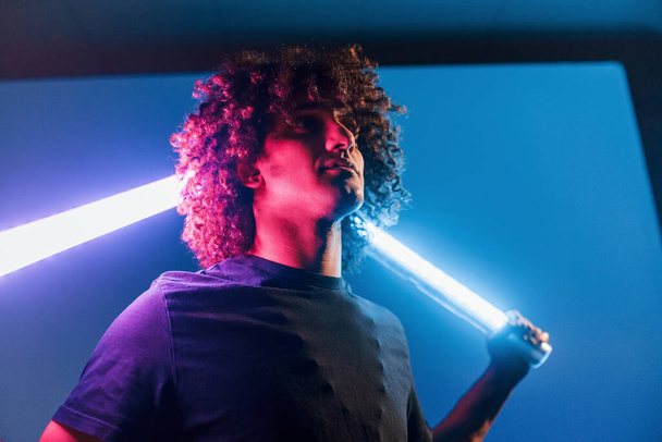 Holds lighting equipment. Young beautiful man with curly hair is indoors in the studio with neon lighting. - Foto, Imagem