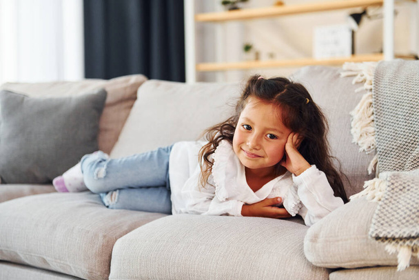 Laying down on the sofa. Little girl in casual clothes is at home alone. - Foto, imagen