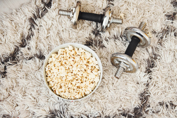 Top view of popcorn in white plate and dumbbells that lying down on the floor. - Φωτογραφία, εικόνα