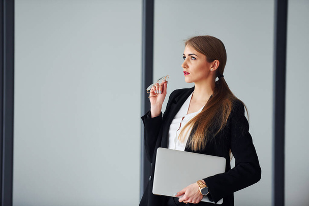 Standing against grey background. Young adult woman in formal clothes is indoors in the office. - Foto, Bild
