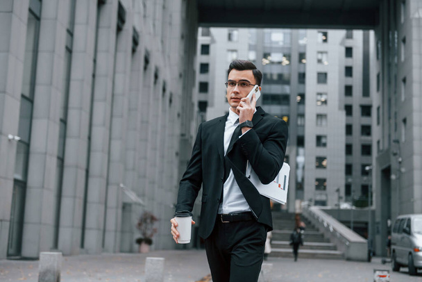 Walking and talking by phone. Businessman in black suit and tie is outdoors in the city. - Foto, Imagem