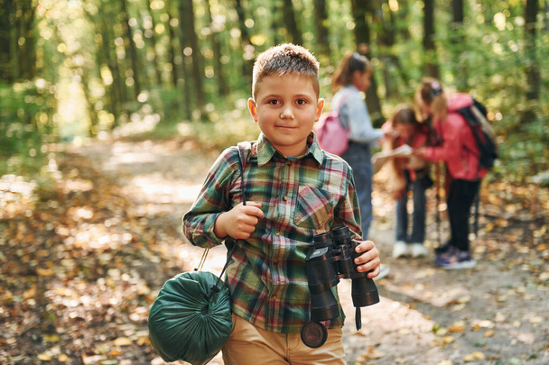 Boy with binoculars standing in front of his friends. Kids in green forest at summer daytime together. - Fotó, kép