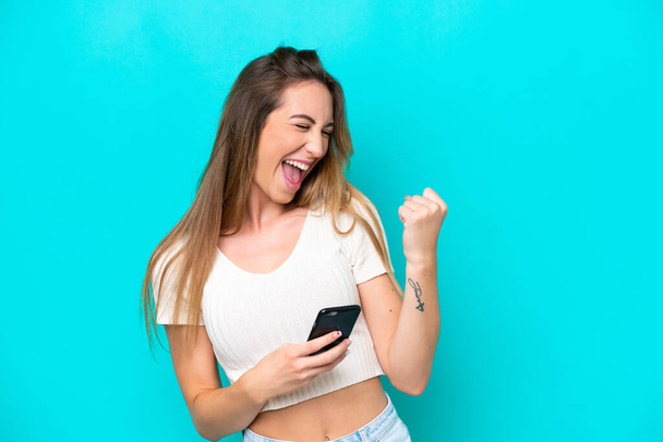 Young caucasian woman isolated on blue background with phone in victory position - Foto, imagen