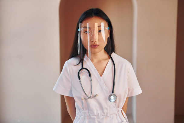In white coat and with stethoscope. Young serious asian woman standing indoors. - Zdjęcie, obraz