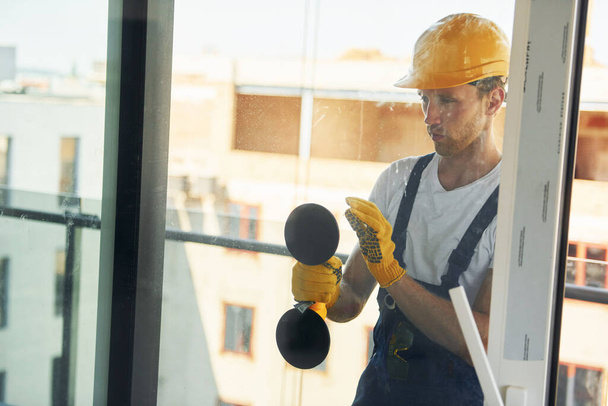 Using tool on glass. Young man working in uniform at construction at daytime. - 写真・画像