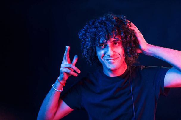 With headphones. Young beautiful man with curly hair is indoors in the studio with neon lighting. - Photo, Image