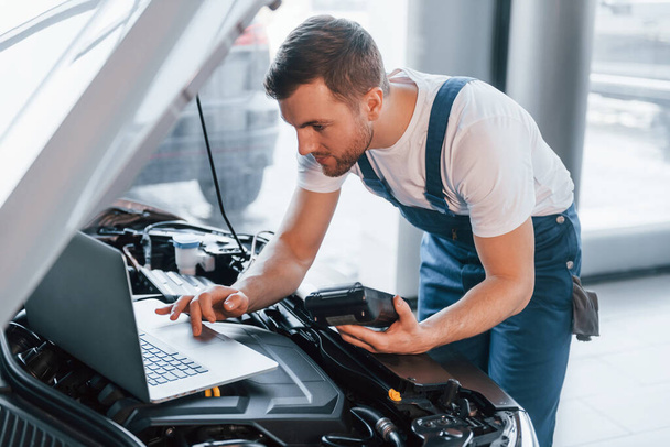 Uses laptop. Young man in white shirt and blue uniform repairs automobile. - Foto, Bild