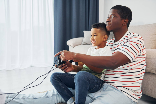 Using joysticks to play video game. African american father with his young son at home. - Foto, afbeelding