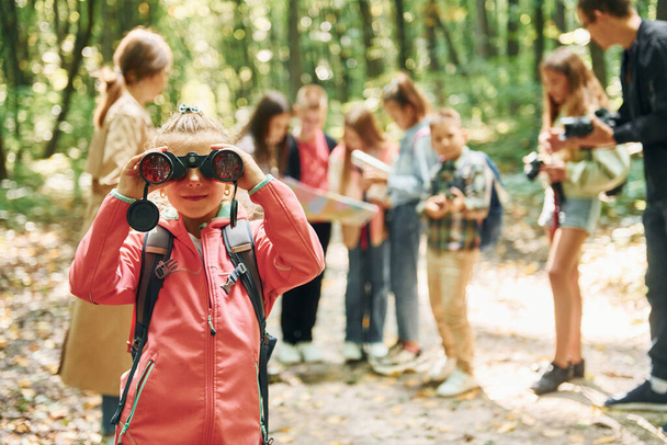 Girl looking into binoculars. Kids in green forest at summer daytime together. - Foto, afbeelding