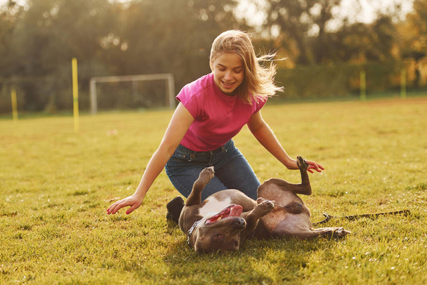 Playing on the ground. Woman in casual clothes is with pit bull outdoors. - Photo, Image