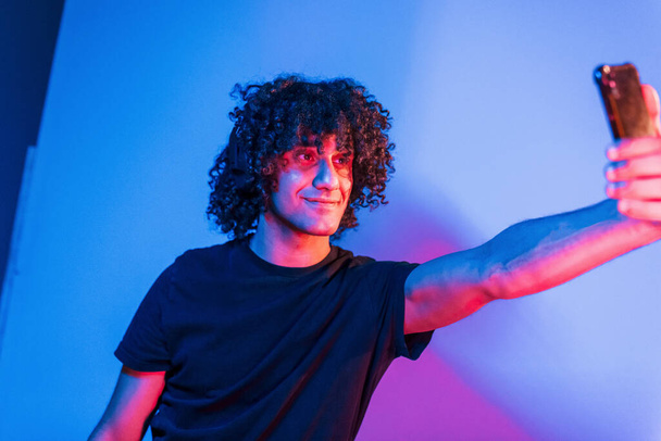 Takes selfie. Young beautiful man with curly hair is indoors in the studio with neon lighting. - Photo, image
