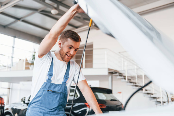 Young man in white shirt and blue uniform repairs automobile. - Foto, imagen
