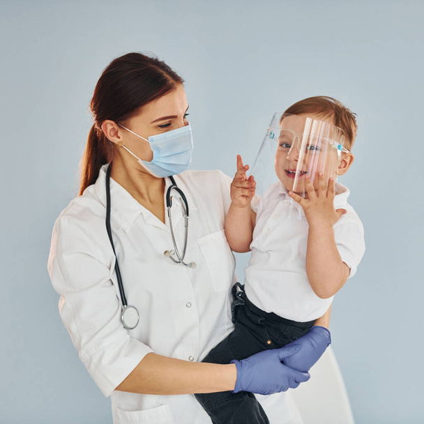 Young nurse in white coat and with stethoscope holds little boy in hands. - Foto, afbeelding