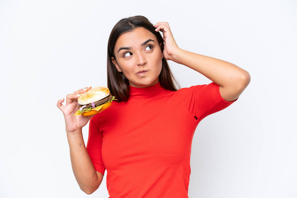 Jovem caucasiana segurando um hambúrguer isolado em fundo branco com dúvidas e com expressão facial confusa - Foto, Imagem