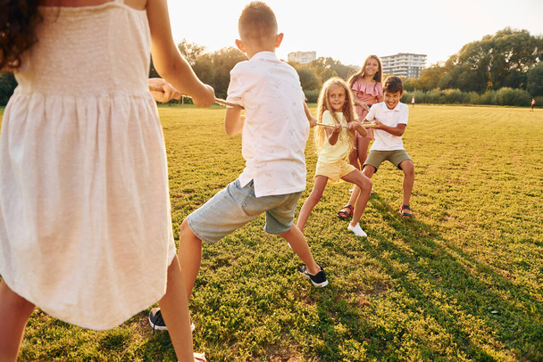 Playing tug of war. Group of happy kids is outdoors on the sportive field at daytime. - Φωτογραφία, εικόνα