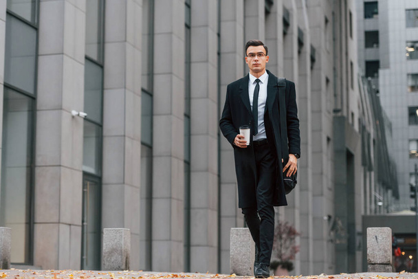 With cup of drink. Businessman in black suit and tie is outdoors in the city. - Foto, Imagen