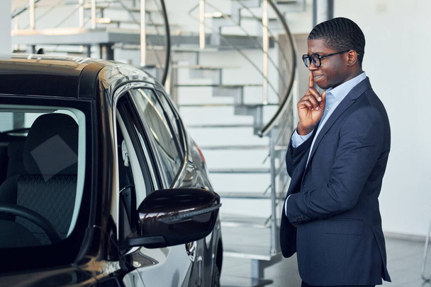 Standing near car. Young african american businessman in black suit is the autosalon. - Foto, immagini
