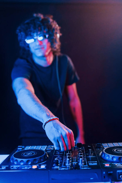 Man with curly hair using DJ equipment and standing in the dark neon lighted room. - Φωτογραφία, εικόνα