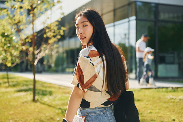 Student walks near modern building. Young asian woman is outdoors at daytime. - Foto, imagen