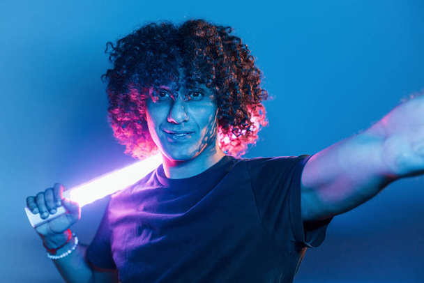 Holds lighting equipment. Young beautiful man with curly hair is indoors in the studio with neon lighting. - Photo, image