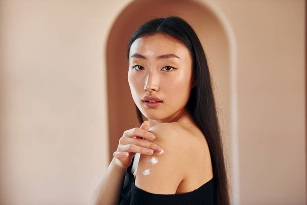 Uses beauty cream. Young serious asian woman standing indoors. - Valokuva, kuva
