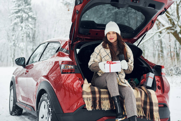 Holding gift box. Beautiful young woman is outdoors near her red automobile at winter time. - 写真・画像
