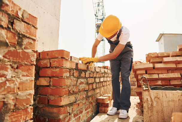 Using bricks. Young construction worker in uniform is busy at the unfinished building. - Fotó, kép