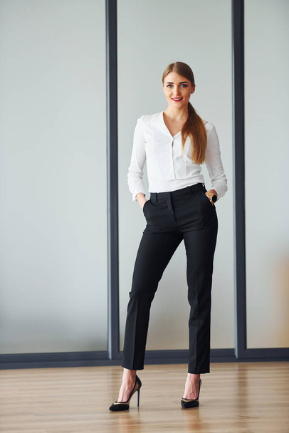 Standing against grey background. Young adult woman in formal clothes is indoors in the office. - Foto, imagen