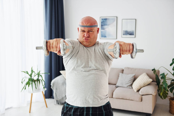 Uses dumbbells. Funny overweight man in casual clothes is indoors at home. - Foto, afbeelding