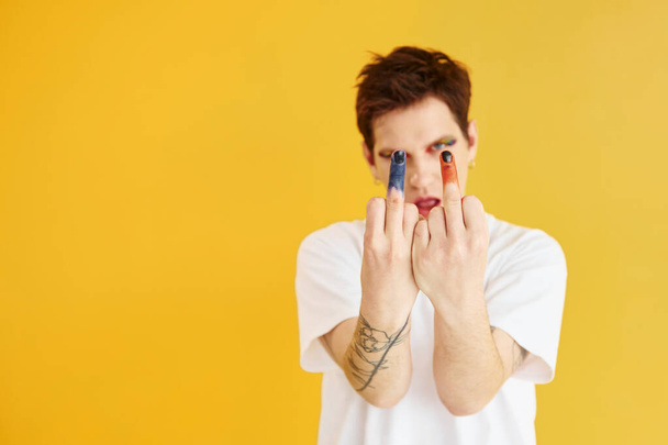 Young gay man is standing in the studio and posing for a camera. Rainbow make up. - 写真・画像