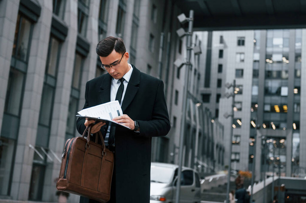 Holding documents. Businessman in black suit and tie is outdoors in the city. - Photo, image