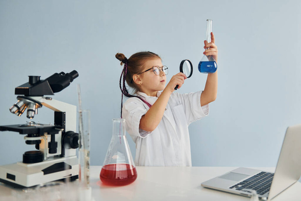 Holds magnifying glass. Little girl in coat playing a scientist in lab by using equipment. - Photo, Image