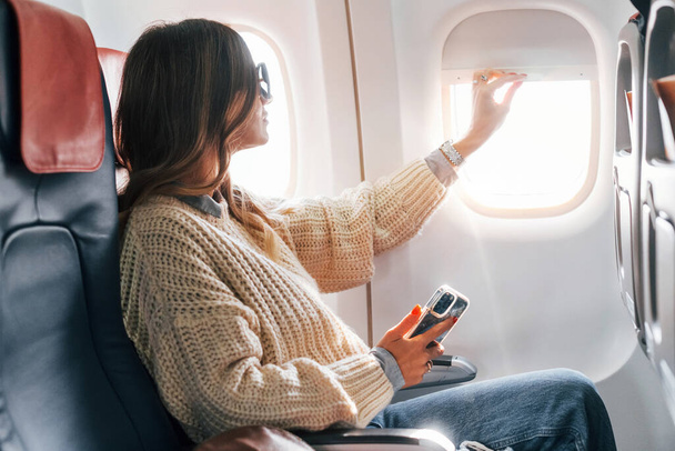 Sitting with phone in hands. Young female passanger in casual clothes is in the plane. - Φωτογραφία, εικόνα