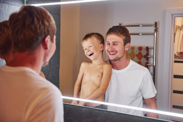 Looking in the mirror in bathroom. Father and son is indoors at home together. - Fotó, kép
