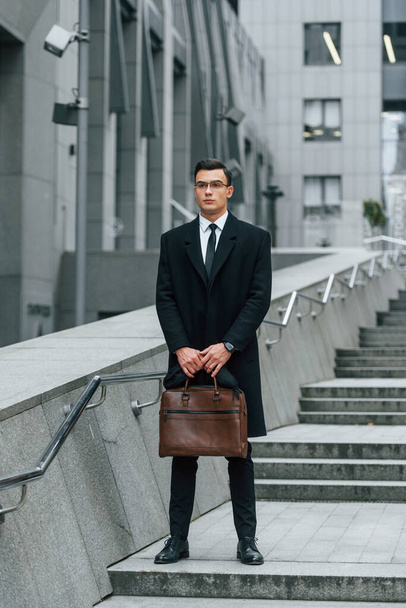 Holding brown bag in hands. Businessman in black suit and tie is outdoors in the city. - Foto, immagini
