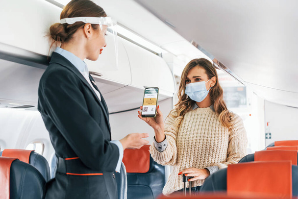 Showing COVID-19 vaccine sertificate to stewardess. Young female passanger in casual clothes is in the plane. - Photo, image