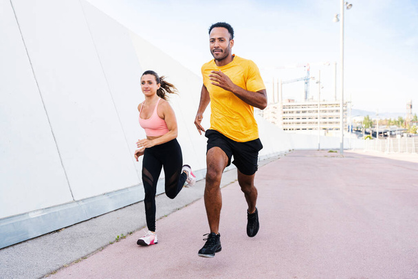 Interracial sportive couple running outdoors - Multiracial couple of runners wearing sportswear and doing functional workout outdoors to strenght body muscles, core abs, stamina and cardio - Foto, immagini