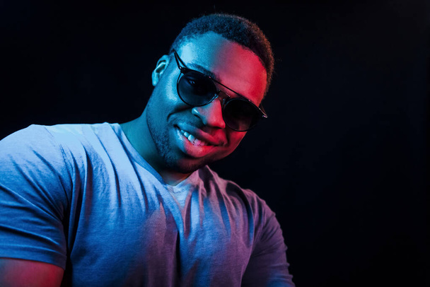 With sunglasses. Futuristic neon lighting. Young african american man in the studio. - 写真・画像