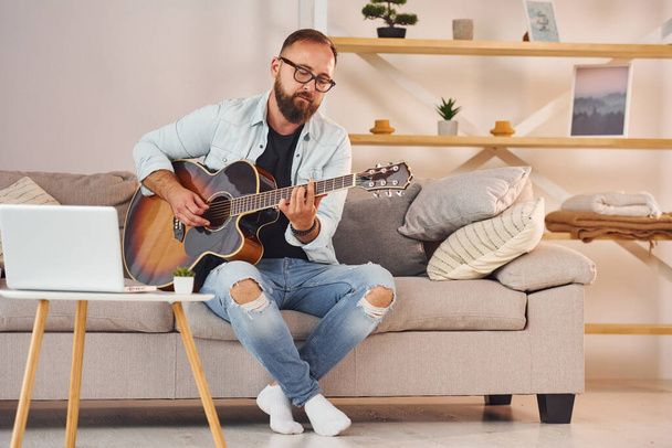 Sitting on the sofa. Man in casual clothes and with acoustic guitar is indoors. - Valokuva, kuva