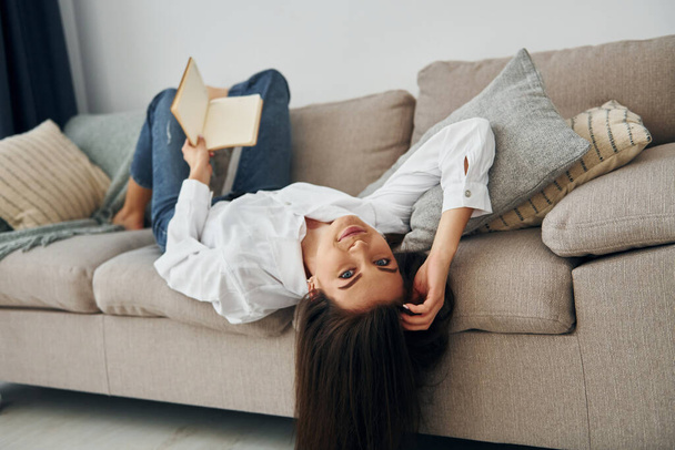 With book. Beautiful young woman in jeans and white shirt is at home. - Foto, Bild