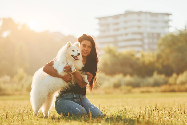 Building at background. Woman with her dog is having fun on the field at sunny daytime. - Foto, imagen
